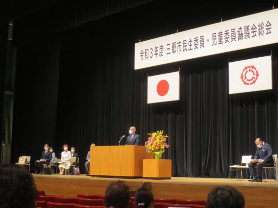 「令和3年度三郷市民生委員・児童委員協議会総会」と書かれた横断幕と国旗、三郷市章が掲げられた会場で三郷市長が立って挨拶をし、関係者の方々が座って聞いている写真