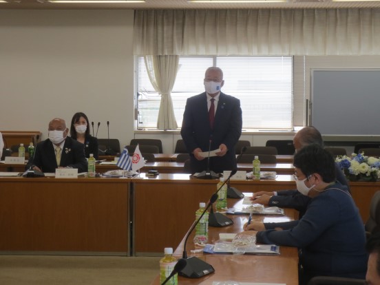 名前のプレートが設置されている席を立って挨拶している三郷市長と、席に座って聞いている関係者の方々の写真
