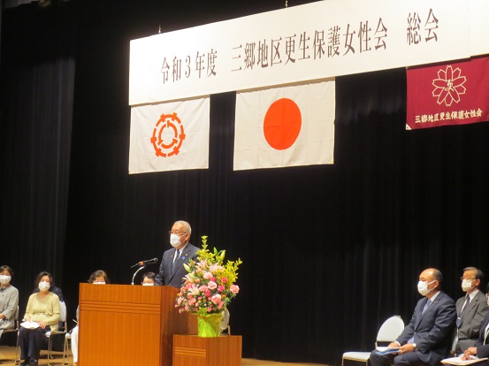 「令和3年度三郷地区更生保護女性会総会」と書かれた横断幕と三郷市章、国旗、三郷地区更生保護女性会の旗が掲げられた会場で、演台に立って挨拶をしている三郷市長と、脇に関係者の方々がパイプ椅子に座って聞いている写真
