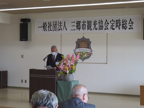 「一般社団法人三郷市観光協会定時総会」と書かれた横断幕と三郷市観光協会会員証マークの前で挨拶をする三郷市長と席に座って聞いている参加者の方々の写真