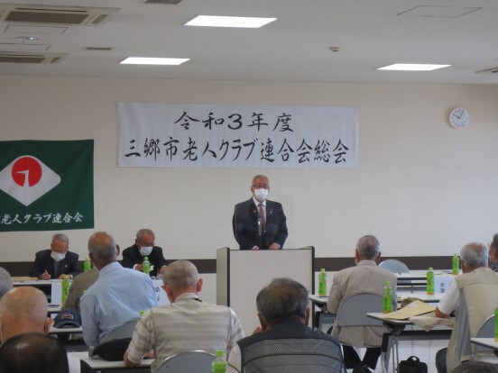 「令和3年度三郷市老人クラブ連合会総会」と書かれた横断幕と老人クラブ連合会の旗と壁に時計が設置された会場で、演台に立って挨拶する三郷市長と2名の関係者、たくさんの参加者の方々が席に座って聞いている写真