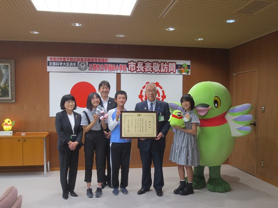 「令和3年度子供の読書活動優秀実践校 文部科学大臣表彰 三郷市立早稲田小学校市長表敬訪問」と書かれたプレートと、国旗、三郷市章が壁に掛けられた部屋で、3人の早稲田小学校の児童、2人の関係者、三郷市長、かいちゃんとつぶちゃんが一緒に記念撮影をしている写真