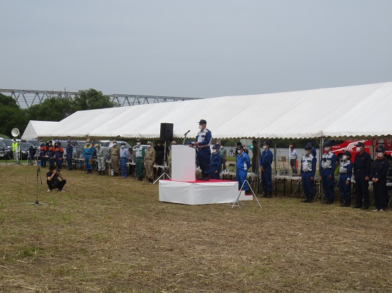 白いイベントテントが設置された会場で三郷市長が演壇に上がり挨拶をし、関係者の方々がイベントテントの前に並んで立って聞いている写真