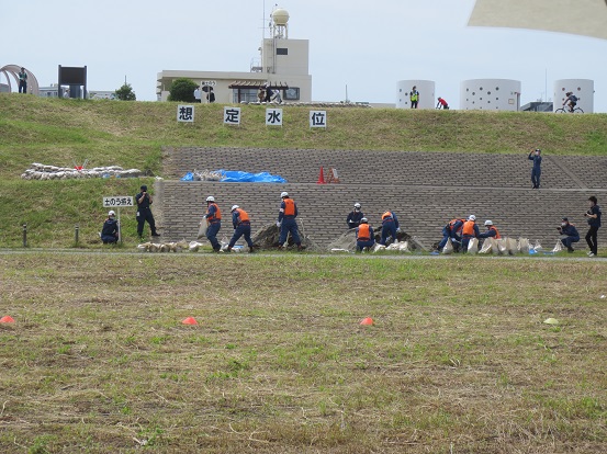 土手の上部に「想定水位」と書かれたプレートが設置され、土手の下部で消防隊員の方々が土のうを並べ消防水防工法訓練を行っている写真