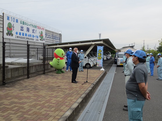 「第10回公園のじゃぐち点検プロジェクト出陣式」と書かれたプレートの前でスタンドマイクの前に立って挨拶をする三郷市長と、かいちゃん、関係者の方々が立って聞いている写真