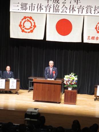 「平成27年三郷市体育協会体育賞授与式」と書かれた横断幕と国旗と三郷市旗が飾られたステージの演壇で挨拶を行っている市長の写真