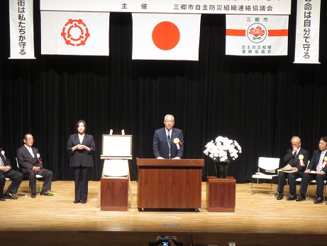 横断幕と国旗や三郷市旗などが飾られたステージの演台で挨拶を行っている市長の写真