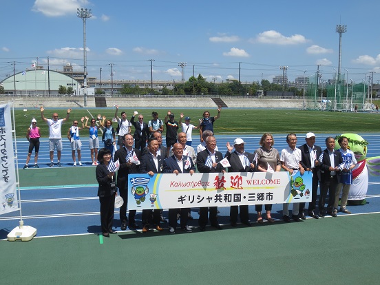 三郷市長、かいちゃん、関係者の方々が間隔を開けて前列と後列に並び、前列は「歓迎ギリシャ共和国・三郷市」と書かれたプレートを一緒に持ち、後列は手を上げて記念撮影をしている写真