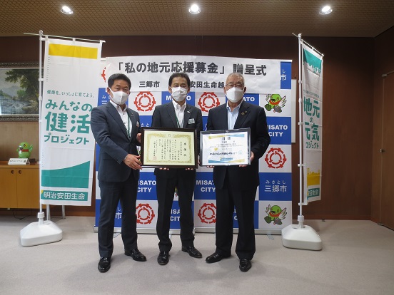 「「私の地元応援募金」贈呈式」と書かれたバックボード、「みんなの健活プロジェクト」と書かれた旗の前で、明治安田生命保険相互会社越谷支社常峰越谷支社長（左）、楠田三郷営業部長（中央）が感謝状を持ち、三郷市長（右）と一緒に記念撮影をしている写真