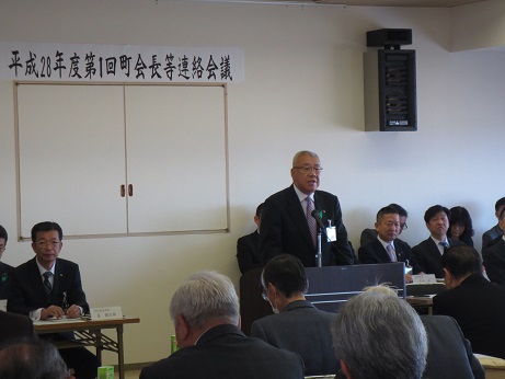 「平成28年度第1回町会長等連絡会議」と書かれた横幕が壁に貼られた室内で、両端の席に座っている関係者と参加者が向かい合って座り、中央の演壇で挨拶を行っている市長の話を聞いている写真