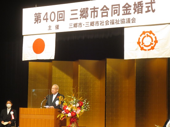 「第40回三郷市合同金婚式」と書かれたプレートと国旗、三郷市章が掲げられた会場の金屏風の前で立って挨拶をしている三郷市長と左脇に関係者の男性が座っている写真