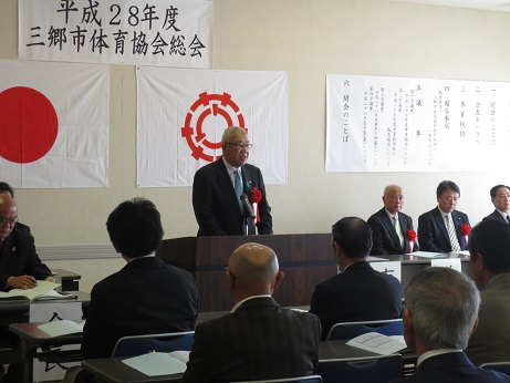 「平成28年度三郷市体育協会総会」と書かれた横幕と国旗と三郷市旗が飾られた室内中央の演壇に立ち挨拶を行っている市長の話を聞いている参加者の後ろ姿の写真
