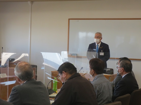前方にホワイトボードが設置された会議室で、自席を立って挨拶する三郷市長とパーテンションで区切られた席に座って聞いている関係者の方々の写真