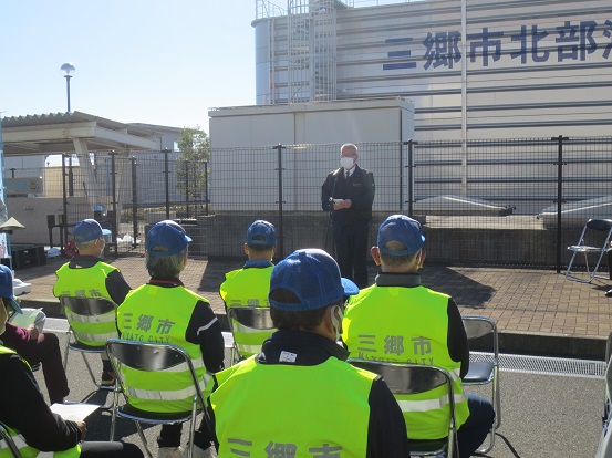 晴天の空の下、スタンドマイクの前に立って挨拶をする三郷市長と蛍光の黄色の「三郷市」と書かれたベストを着た方々がパイプ椅子に座って聞いている写真