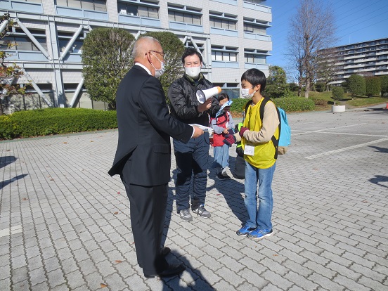 市役所南側駐車場で男の子が三郷市長へ報告書を手渡しし、その横で男性が拡声器を持っている写真
