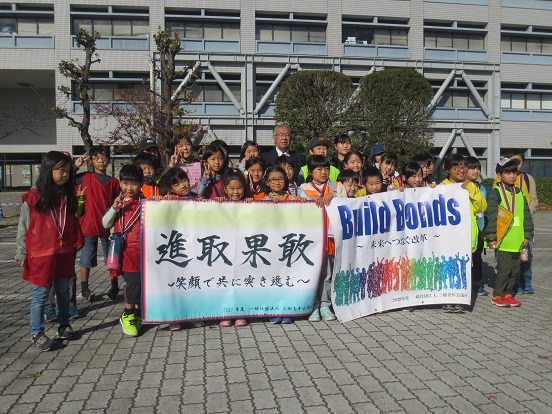 「進取果敢 笑顔で共に突き進む」「Build Bonds 未来へつなぐ改革」と書かれた2つの横断幕をたくさんの子どもたちが持ち、三郷市長と一緒に記念撮影をしている写真
