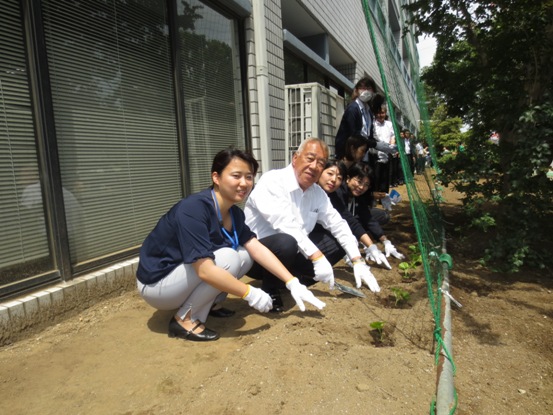 軍手をした数名の職員と市長が建物前の土にゴーヤの苗を植えてカメラ目線で微笑んでいる写真