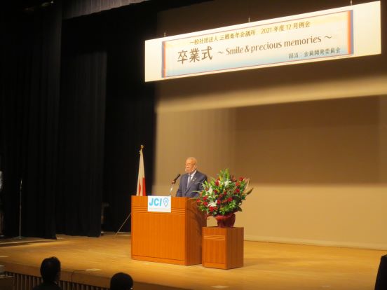 「一般社団法人三郷青年会議所2021年度12月例会 卒業式 Smile＆precious memories」と書かれたプレートが掲げられた会場で、三郷市長が演台に立ち挨拶をしている写真