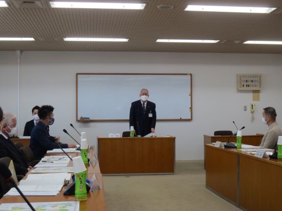 前方にホワイトボードが設置された会議室で、自席を立って挨拶する三郷市長と向かい合わせに設置されている席に座り聞いている関係者の方々の写真