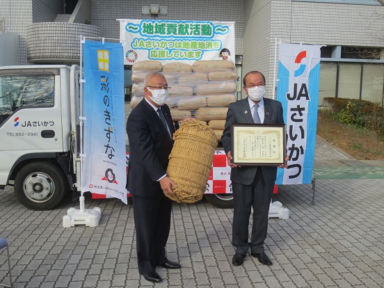 トラックの荷台にたくさんのお米が積み上げられ、脇に「JAさいかつ」「彩のきずな」と書かれた2つの旗が飾られた会場で、三郷市長が米俵を持ちJAさいかつの男性が感謝状を持って記念撮影をしている写真