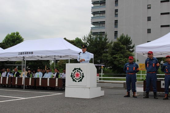 駐車場に設置されたテントの中央の演壇に立つ市長が挨拶を行っている様子の写真