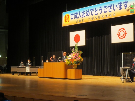 「祝 ご成人おめでとうございます」と書かれたプレートと国旗、三郷市章が掲げられた会場で、演台に立って挨拶をしている三郷市長と手話通訳の女性、関係者の方々が席に座っている写真