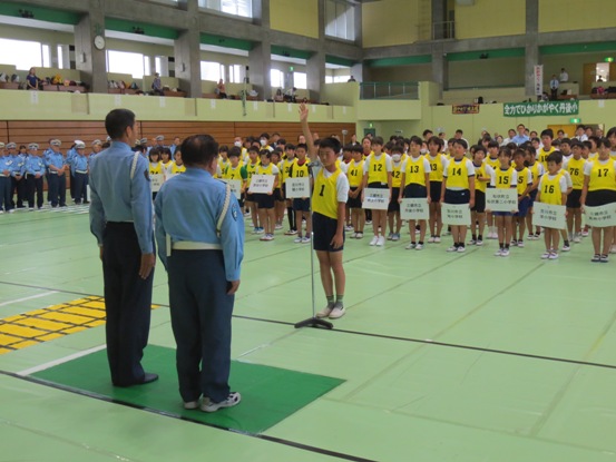 黄色のゼッケンをつけた子ども達が整列した体育館で、警察官のかたに向かって1名の男の子が代表で選手宣誓をしている写真