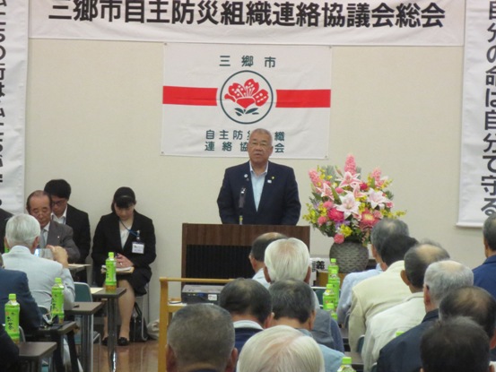 壁に「三郷市自主防災組織連絡協議会総会」と書かれた紙が貼ってある部屋で、多くの参加者が椅子に着席しているなか、正面の演台に立って話をしている市長の写真
