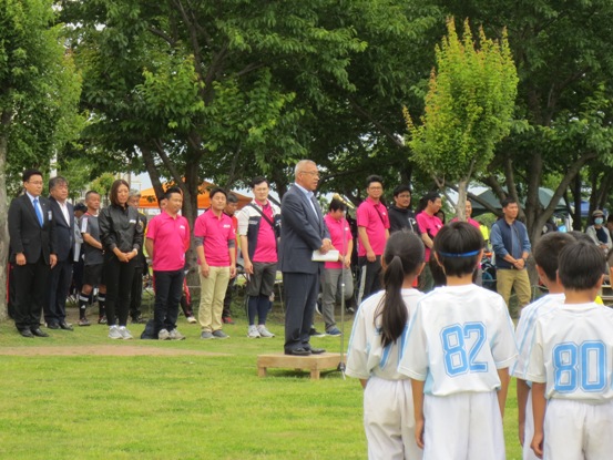 芝生の広がる広場で、関係者や児童達が台の上に立って話をしている市長を見ている写真