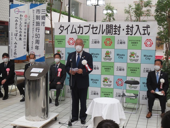 「タイムカプセル開封・封入式」と書かれた三郷市のバックボードの前で三郷市長が立って挨拶をし、関係者の方々がパイプ椅子に座っている写真