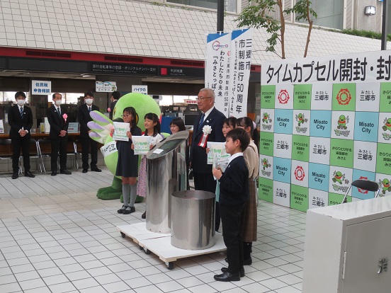 「タイムカプセル開封・封入式」と書かれた三郷市のバックボードの前で、かいちゃん、6人の子どもたち、三郷市長がタイムカプセルを囲んで一緒に記念撮影をしている写真