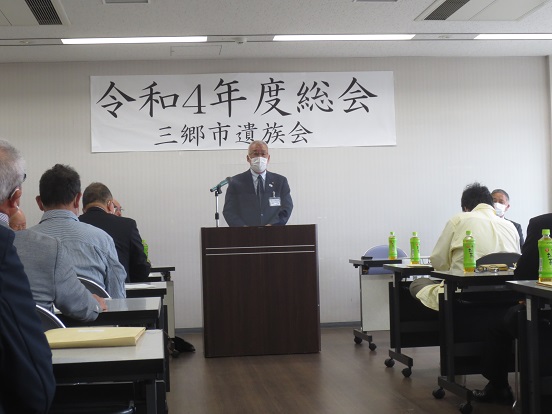 「令和4年度総会三郷市遺族会」と書かれた横断幕が掲げられた会場で、三郷市長が演台に立ち挨拶をし、長机と椅子が設置された席に座って聞いている参加者の方々の写真