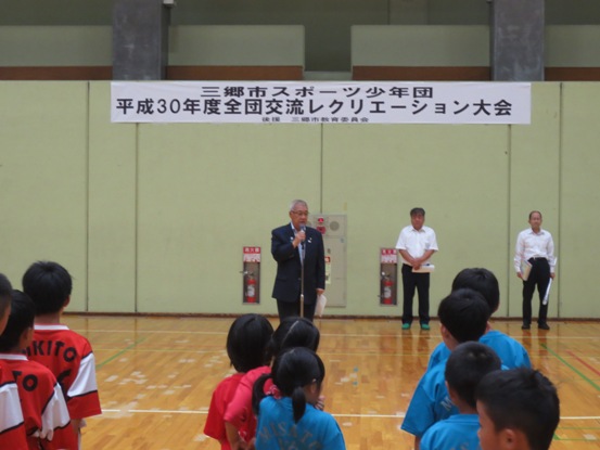 「三郷市スポーツ少年団 平成30年度全団交流レクリエーション大会」の紙が貼られた体育館で、整列している子どもたちの正面に立って話をしている市長の写真