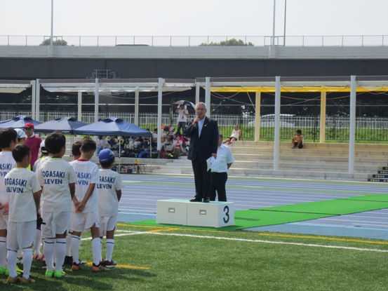 三郷市陸上競技場内の芝生上にたくさんの子ども達が整列しているなか、正面に用意された台の上に立って話をしている市長の写真