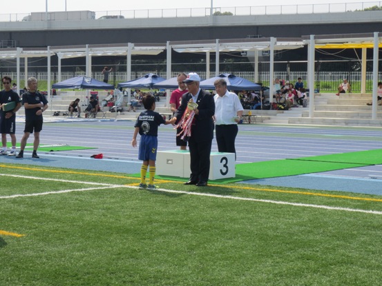 三郷市陸上競技場の芝生の上で、一名の男児が市長へ優勝カップを返還している写真