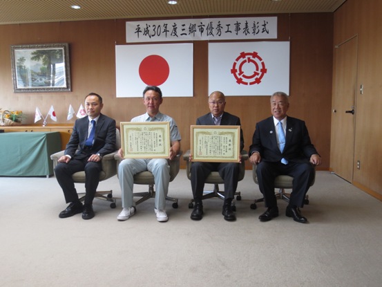 「平成30年度三郷市優秀工事表彰式」の紙が設置された部屋で、市長と関係者の間に座っている2名の受賞者が表彰状を持っている写真