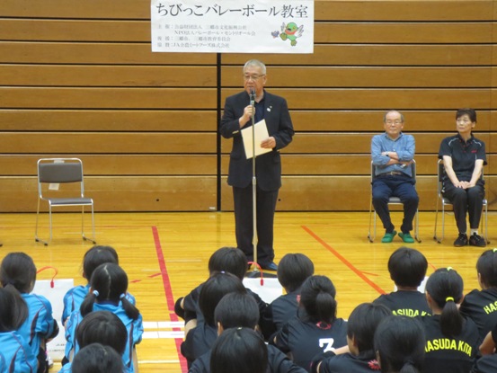 「ちびっこバレーボール教室」の紙が貼られた体育館で、チームごとに並んで座る子どもたちの正面に立って話をしている市長の写真