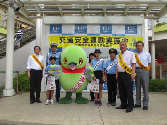 「交通安全運動実施中」の横断幕が設置された施設のステージ上で、一日署長の子どもたちや関係者たち、かいちゃんと一緒に記念撮影をしている写真