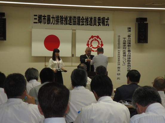 「三郷市暴力排除推進協議会推進員委嘱式」と書かれた紙が設置されている部屋で、多くの関係者たちが座って見守る中、部屋の正面で市長から一名の男性へ委嘱書が交付されている様子の写真