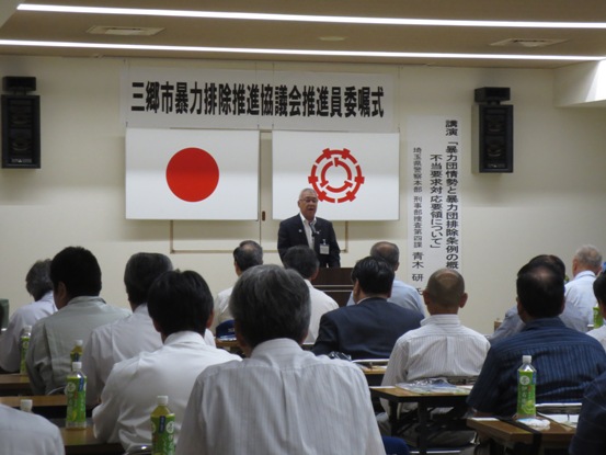 「三郷市暴力排除推進協議会推進員委嘱式」と書かれた紙が設置され、多くの関係者たちが座っている部屋で、正面に立って話をしている市長の写真