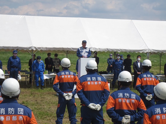 白いテントが建てられた会場の前方で三郷市長が演台に立って訓示を述べており、オレンジとネイビーの制服を着た消防団の方々が整列し手を後ろに組んで聞いている写真