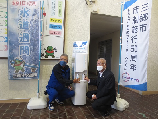 関係者の男性と三郷市長が寄贈いただいたマイボトル用給水機と一緒にしゃがんで記念撮影をしている写真