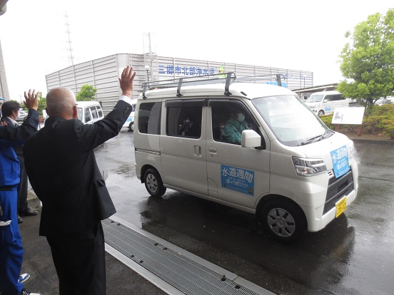 「水道週間」と書かれたプレートを付けた車で蛇口点検に出発する関係者の方々を手を振って見送っている三郷市長の写真