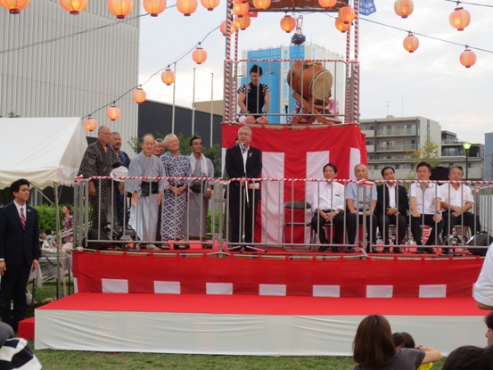 芝生に紅白の横断幕で囲われた特設ステージが置かれ、一番上の段のステージには太鼓と一名の男性、2段目には着物やスーツ姿の関係者が控えるなか、中心に立つ市長が挨拶をしている写真