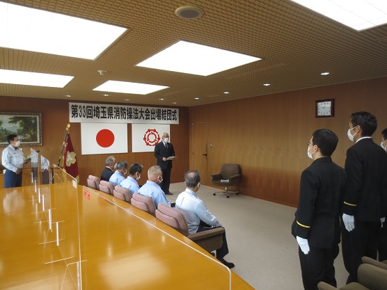 「第33回埼玉県消防操法大会出場結団式」と書かれた横断幕と国旗、三郷市章が壁に掛けられた部屋の前方で立って挨拶する三郷市長と、座って聞いている関係者の方々、直立姿勢をとっている黒い制服を着た男性の方々の写真