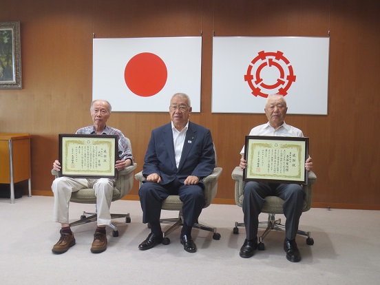 日本国旗と三郷市旗の前に設置された椅子に座って、感謝状を持っている斎藤誓任様と大野勇様、中央に座る木津市長の記念写真