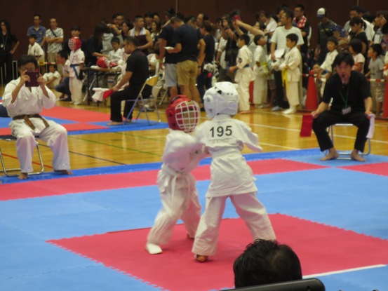 関係者が見守る中、白の道着やヘッドガードを着用した2名の子どもが試合を行っている写真