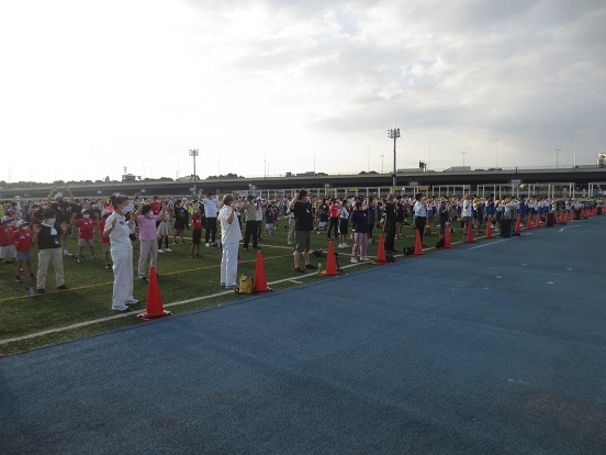 三郷市陸上競技場の芝生上で様々な年代のかたが等間隔に並んで体操をしている様子の写真