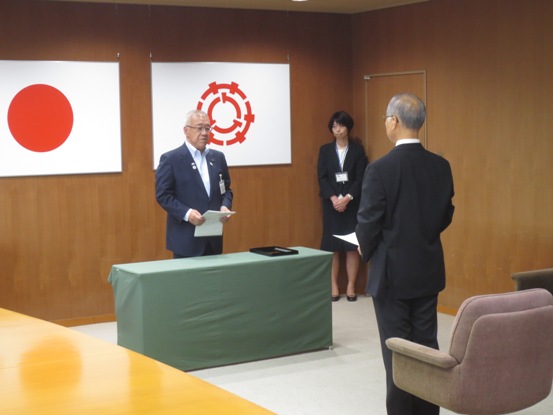 日本国旗や三郷市章が掲げられている部屋で、秘書の女性が見守る中、市長と男性が向かい合って任命式を行っている様子の写真