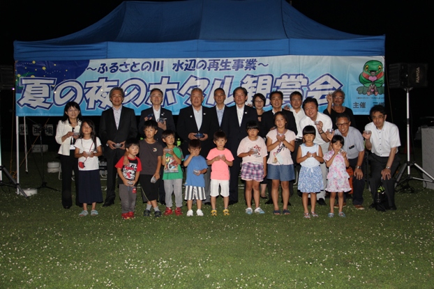 「夏の夜のホタル観賞会」の横断幕の前に立って、子どもたちや関係者たち合わせて総勢22名の参加者と一緒に並んで記念撮影をしている市長の写真